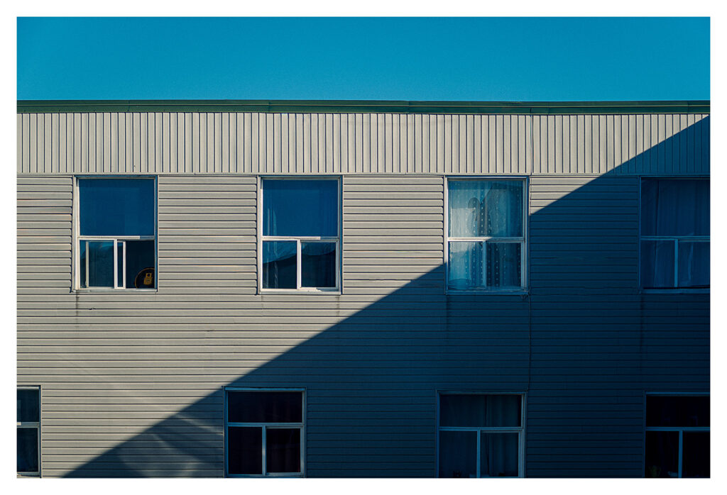 Un bâtiment de deux étages avec un revêtement en tôle sous un ciel bleu clair. Plusieurs fenêtres sont visibles, certaines ouvertes et d'autres avec des rideaux. Une ombre nette traverse le bâtiment en diagonale, ajoutant un contraste entre les sections ensoleillées et ombragées.
