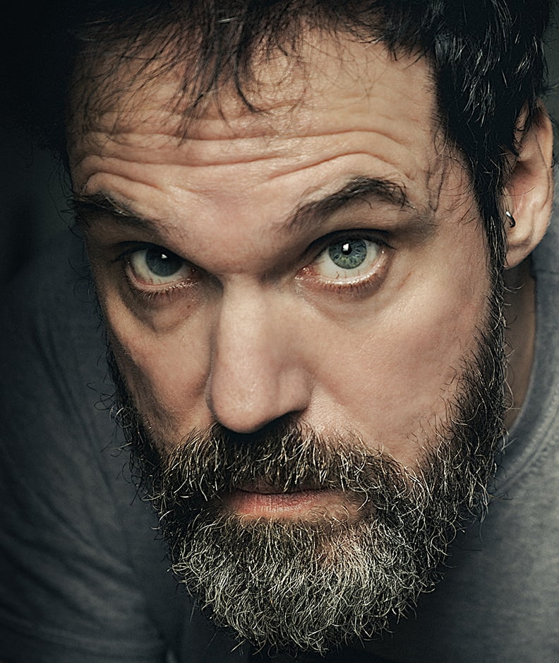 Image en gros plan d'un homme avec une barbe et une moustache épaisses, des yeux bleus intenses et un sourcil légèrement plissé. Il a les cheveux courts et foncés avec des reflets gris et porte une chemise grise. L’arrière-plan est sombre, attirant l’attention sur son visage.