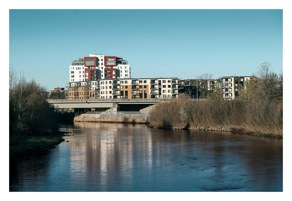 Un complexe d'appartements moderne de plusieurs étages aux façades variées se dresse au bord d'une rivière calme, reflétant le ciel bleu clair. Les bâtiments sont entourés d'arbres dénudés et de végétation, avec un pont visible au premier plan.