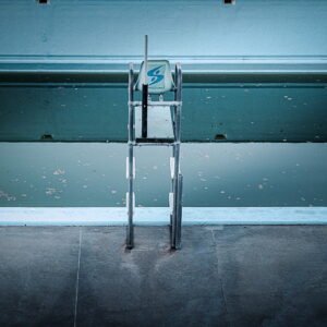 Un bloc de plongée se dresse au bord d’une piscine vide et faiblement éclairée. Les marches métalliques reflètent une lumière bleue tamisée et l’eau de la piscine est calme, avec quelques feuilles flottant à la surface. L'atmosphère générale est calme et légèrement inquiétante.