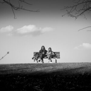 Deux personnes sont assises sur un banc de parc sur une colline herbeuse, avec des nuages épars dans le ciel derrière elles. Tous deux sont occupés avec leur téléphone ou leurs appareils, et des arbres aux branches nues encadrent la photo dans les coins. L'image est en noir et blanc.