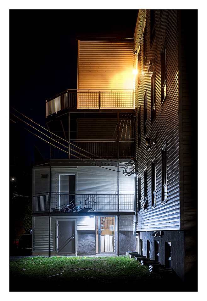 Une vue nocturne de l'extérieur d'un immeuble à plusieurs étages avec balcons et balustrades. Le bâtiment est éclairé par diverses lumières, projetant une lueur chaleureuse sur les balcons supérieurs et une lumière plus froide et vive sur la partie inférieure. Les lignes électriques sont visibles à gauche.