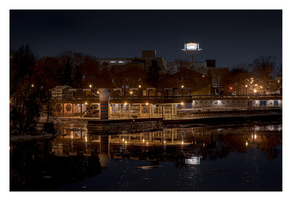 Vue nocturne d'une zone riveraine avec un château d'eau éclairé en arrière-plan. La scène comprend des bâtiments éclairés et leurs reflets dans l'eau calme. Des arbres et autres structures sont visibles, le tout sous un ciel sombre.