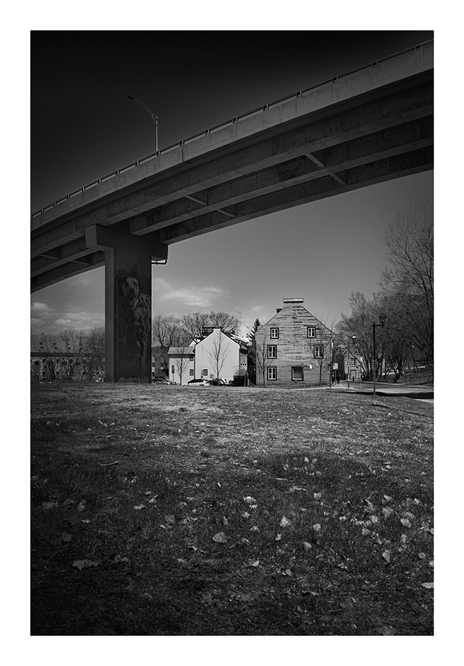 Photographie en noir et blanc d'un viaduc passant sur un sol herbeux parsemé de feuilles mortes. Sous le pont se trouvent quelques bâtiments anciens. L'un des supports du pont présente une peinture murale. Des arbres sans feuilles se trouvent de chaque côté du pont. Le ciel est clair.