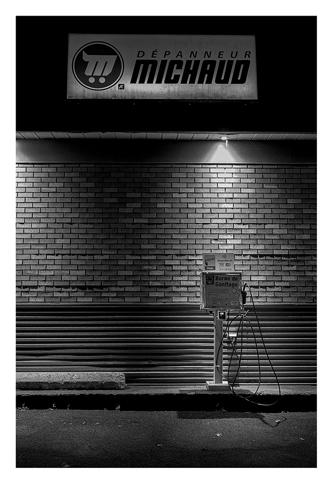 Une photo en noir et blanc d'une enseigne « Dépanneur Michaud » sur un mur de briques. Sous le panneau se trouve une lumière éclairant un distributeur automatique ou une station de recharge avec des tuyaux. La scène globale est faiblement éclairée, créant une atmosphère maussade.