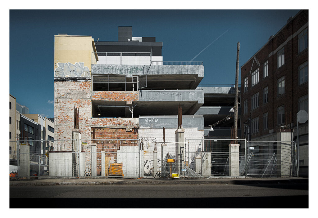 Un bâtiment partiellement démoli avec des murs en briques apparentes, des débris de construction et des structures en béton incomplètes. Le site est entouré d'une clôture métallique et entouré de bâtiments modernes sous un ciel clair et ensoleillé. Des graffitis sont visibles sur certaines surfaces.