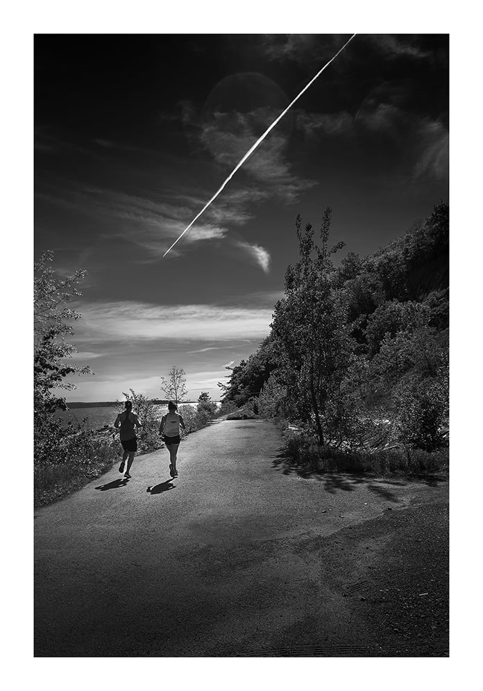 Deux personnes courent sur un chemin pavé entouré d'arbres et de feuillages, sous un ciel clair avec une traînée de condensation proéminente. La scène est en noir et blanc, projetant des contrastes et des ombres spectaculaires.