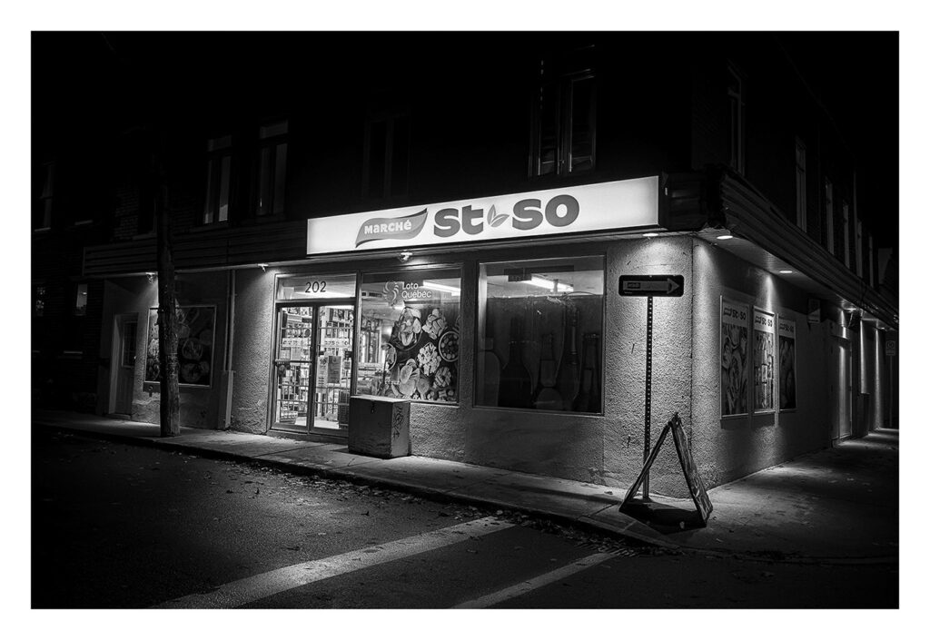Un petit dépanneur nommé « Marché St. So » est illuminé la nuit par une signalisation lumineuse. Les vitrines du magasin présentent divers produits et un panneau de signalisation est visible près de l'entrée. La rue et le trottoir environnants sont sombres et vides.