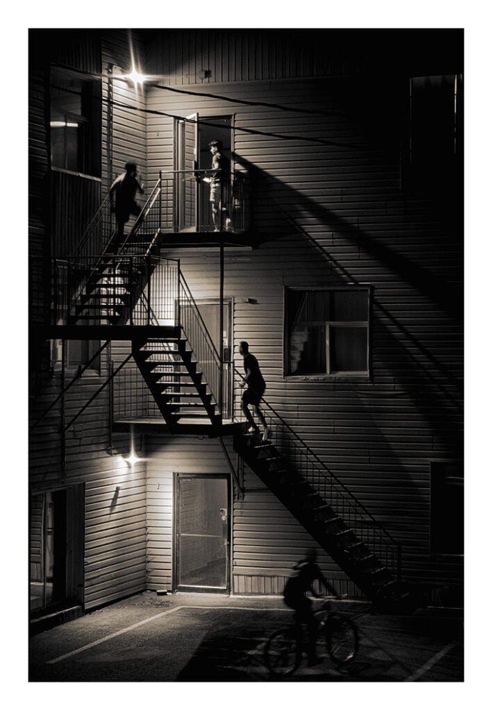 Une photo en noir et blanc montre trois silhouettes sombres sur un escalier extérieur faiblement éclairé d’un immeuble. Un autre personnage sur une bicyclette se déplace au premier plan. La scène est nocturne avec des lumières projetant des ombres dramatiques sur les murs.