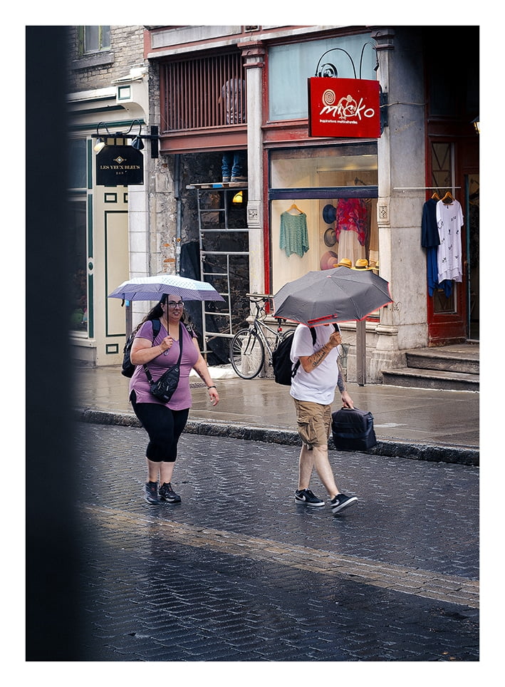 Deux personnes marchent dans une rue pavée sous la pluie. Ils utilisent des parapluies ; une personne tient un parapluie gris tandis que l’autre en tient un transparent. Un vélo est garé devant un magasin de vêtements, avec un panneau rouge au-dessus de la porte et divers vêtements exposés en vitrine.