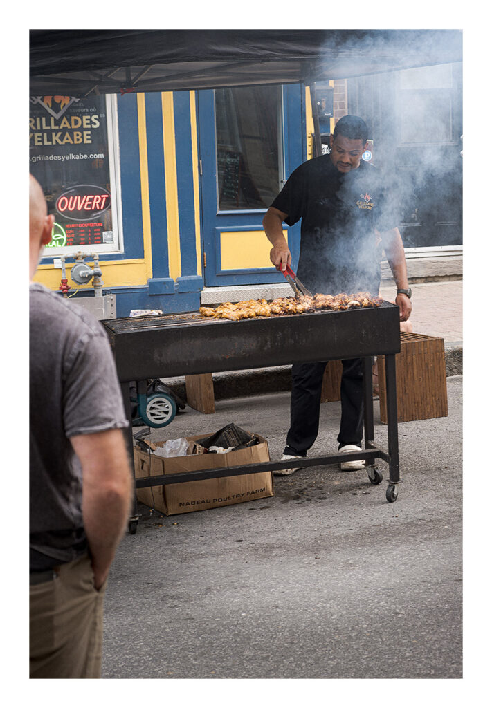 Grillades Un homme fait griller de la viande sur un grand barbecue en plein air sous un auvent dans une rue de la ville. De la fumée s'échappe du gril. Il se tient devant un bâtiment aux accents bleus et jaunes. Deux autres personnes sont partiellement visibles, l'une au premier plan et l'autre à gauche.