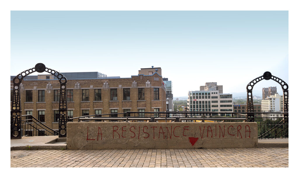 Vue d'une ville avec des immeubles de hauteur moyenne et un grand mur de briques beiges au premier plan. Le mur est orné de graffitis rouges sur lesquels on peut lire « LA RESISTANCE VAINCRA », ce qui signifie « LA RESISTANCE VAINCRERA » en français. Deux portes en fer forgé encadrent le mur.