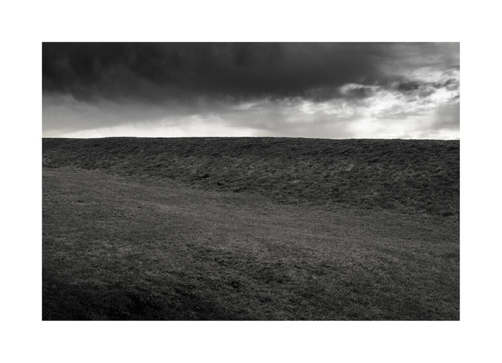 Une photographie en noir et blanc d’un paysage herbeux sous un ciel nuageux. Les nuages sombres se profilent au-dessus d’une colline, projetant une ombre sur le terrain. L’image dégage une atmosphère maussade et dramatique.