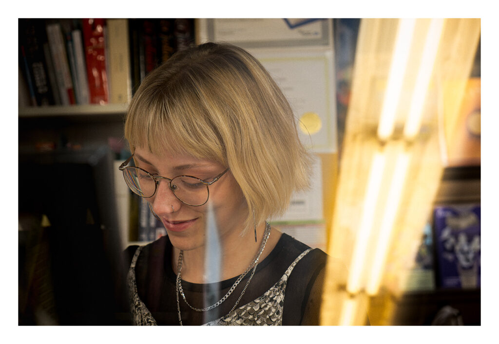 Océane Une personne aux cheveux blonds courts et aux lunettes sourit et regarde vers le bas. Elle porte un haut noir avec un collier en argent. L'arrière-plan comprend des étagères et des certificats, et le premier plan présente des lumières jaunes floues.