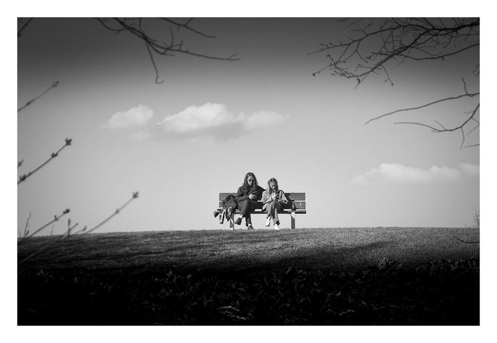 Photo en noir et blanc de deux personnes sur un banc de parc Une photo en noir et blanc de deux personnes assises sur un banc au sommet d’une colline herbeuse. Ils sont engagés dans une conversation avec un minimum de détails en arrière-plan, juste quelques nuages dans le ciel et quelques branches encadrant l'image.