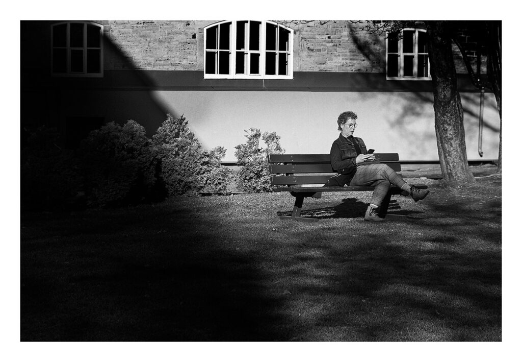 Photo en noir et blanc d'une personne sur un banc dans un parc Une personne est assise seule sur un banc de parc, regardant son smartphone au soleil. Ils sont entourés d’ombres projetées par des arbres et des buissons, avec un bâtiment en brique en arrière-plan. La scène est paisible et tranquille. L'image est en noir et blanc.
