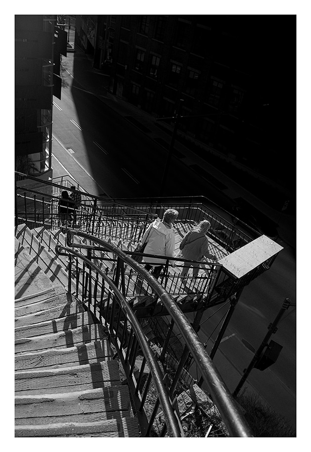 Image en noir et blanc de deux personnes descendant un escalier raide muni de rampes, à côté d’une rue d’une zone urbaine. Les gens sont habillés chaudement et des ombres sont projetées sur les marches et les rampes, suggérant une forte lumière du soleil venant du côté gauche.