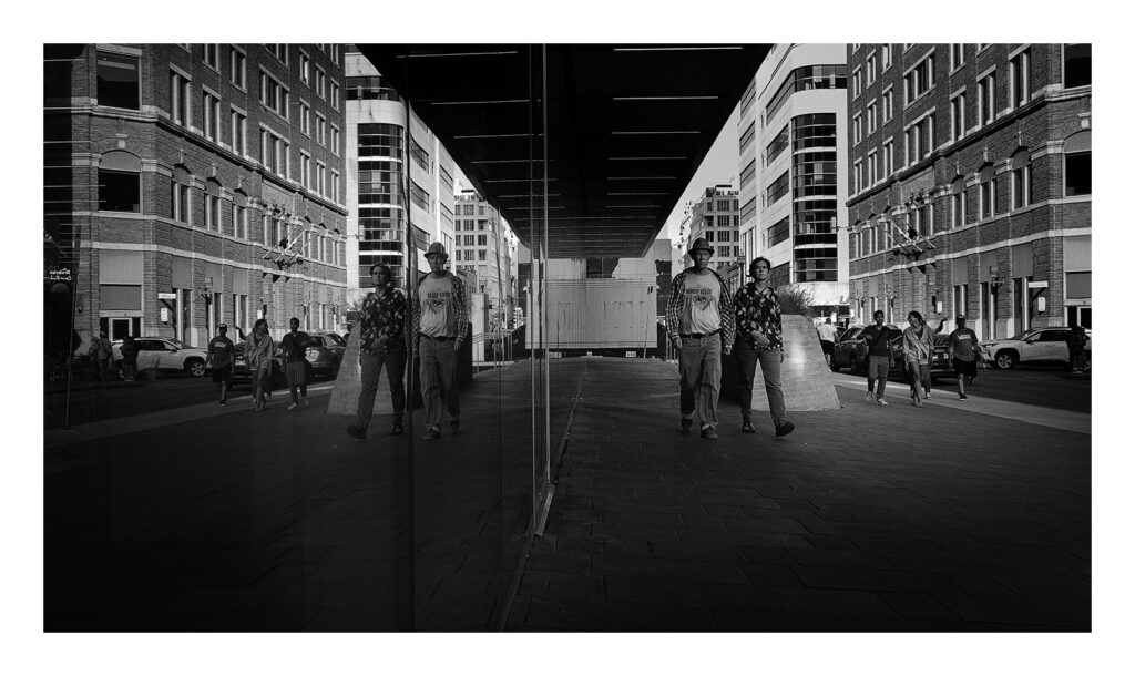 Réflexion Une photographie en noir et blanc capture une scène urbaine animée avec des piétons marchant le long d'un trottoir flanqué de hauts bâtiments. Le centre de l'image présente une surface en verre réfléchissant, créant un effet miroir qui dédouble les personnes et l'architecture.