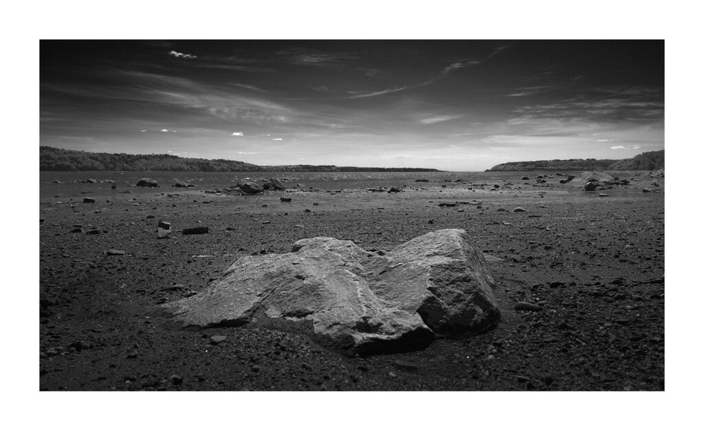 Photographie en noir et blanc d’un paysage aride et rocheux. Plusieurs gros rochers sont disséminés au premier plan avec une vaste étendue vide s'étendant jusqu'à l'horizon. Des collines clairsemées et un ciel partiellement nuageux sont visibles au loin.
