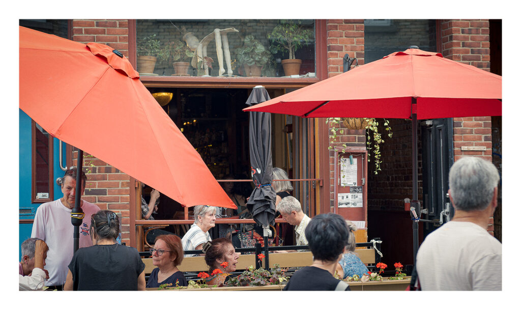 Terrasse rouge d'été Des gens profitent de la nourriture et discutent dans un café en plein air sous de grands parasols rouges. La façade en brique du bâtiment, les plantes en pot et l'atmosphère accueillante sont visibles. Plusieurs clients participent à diverses activités, créant une ambiance animée.