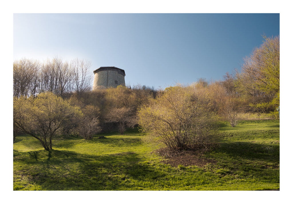 Une tour de pierre se dresse au sommet d’une colline, entourée d’arbres dénudés et de buissons au feuillage clairsemé. Le premier plan présente un terrain verdoyant et herbeux avec des buissons épars. Le ciel au-dessus est clair et bleu, indiquant une journée ensoleillée.