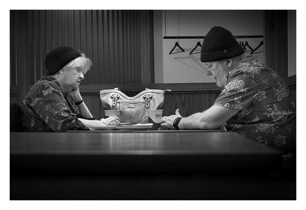deux-personnes-dans-un-cafe-se-font-face Photo en noir et blanc d’un couple de personnes âgées assis à une table dans un restaurant. Tous deux portent des bonnets et des chemises à motifs. L'homme est concentré sur un appareil dans sa main tandis que la femme le regarde pensivement. Un sac à main est accroché à la cloison derrière eux.