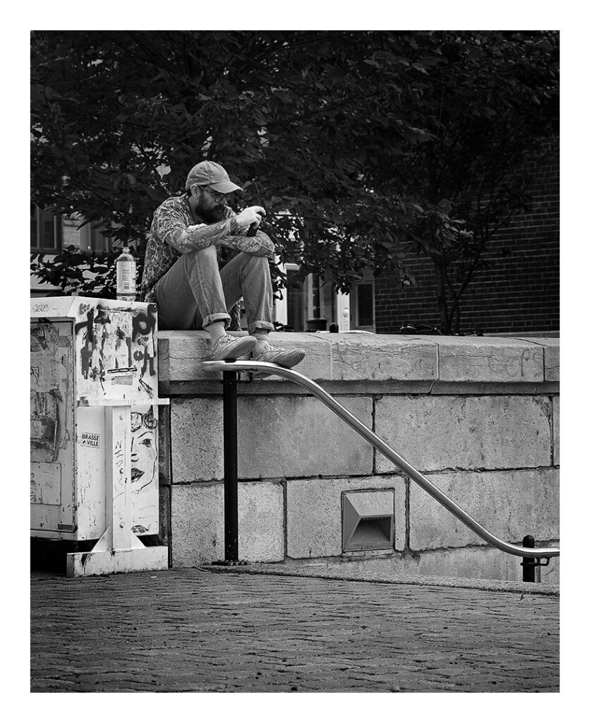 le penseur de l'église Photographie en noir et blanc d'un homme assis sur un rebord de béton, portant une casquette et des vêtements décontractés. Il boit dans une tasse, un bras posé sur son genou. Derrière lui se trouve un arbre et à sa gauche, une structure couverte de graffitis.