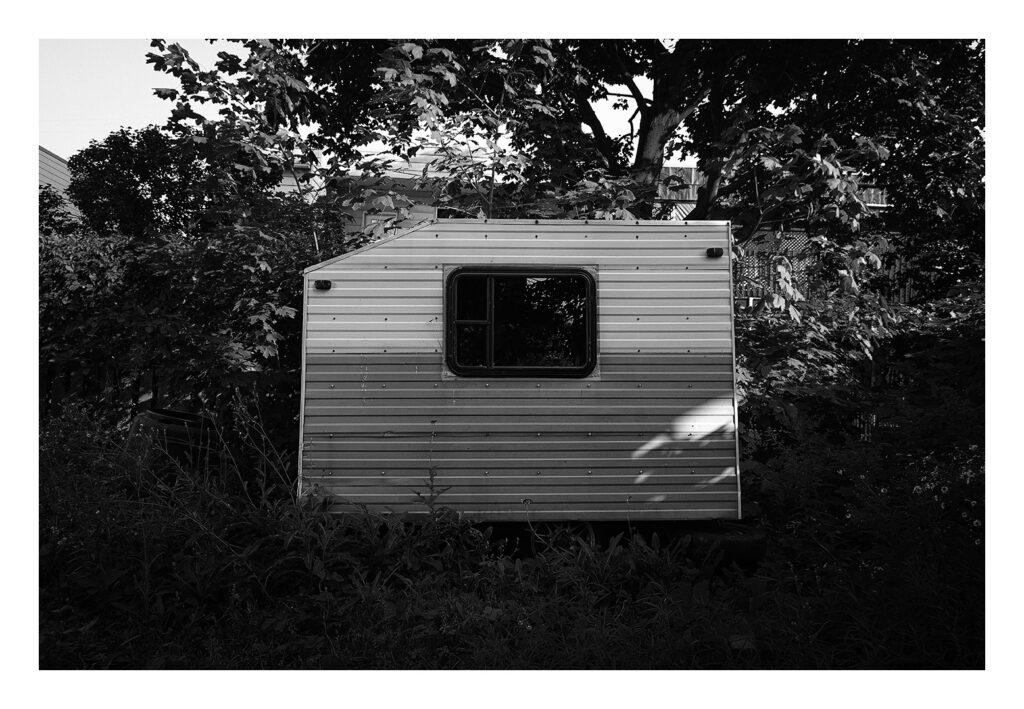 Photographie en noir et blanc d'une petite caravane vintage entourée d'un feuillage dense et d'arbres. La caravane est dotée d'une seule fenêtre carrée et d'un revêtement métallique exposé. La lumière du soleil illumine partiellement la surface, projetant des ombres sur les plantes environnantes.