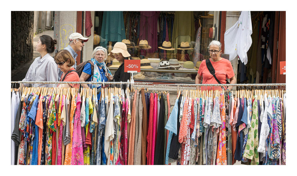 Des vêtements colorés Un groupe de personnes parcourt les rayons de vêtements colorés exposés à l'extérieur d'un magasin. Les présentoirs portent des panneaux indiquant une réduction de 50 %. En arrière-plan, des chapeaux et d'autres articles sont visibles dans la vitrine du magasin. Une femme porte une chemise rouge et des lunettes.